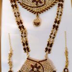 A stunning Rajasthani Royal Gold-Plated Bridal Jewelry Set, featuring a necklace, earrings, and a maang tika, adorned with crimson and ivory gemstones and intricate designs.