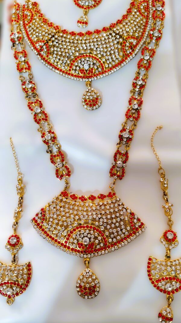 A stunning Rajasthani Royal Gold-Plated Bridal Jewelry Set, featuring a necklace, earrings, and a maang tika, adorned with red and white gemstones and intricate designs.